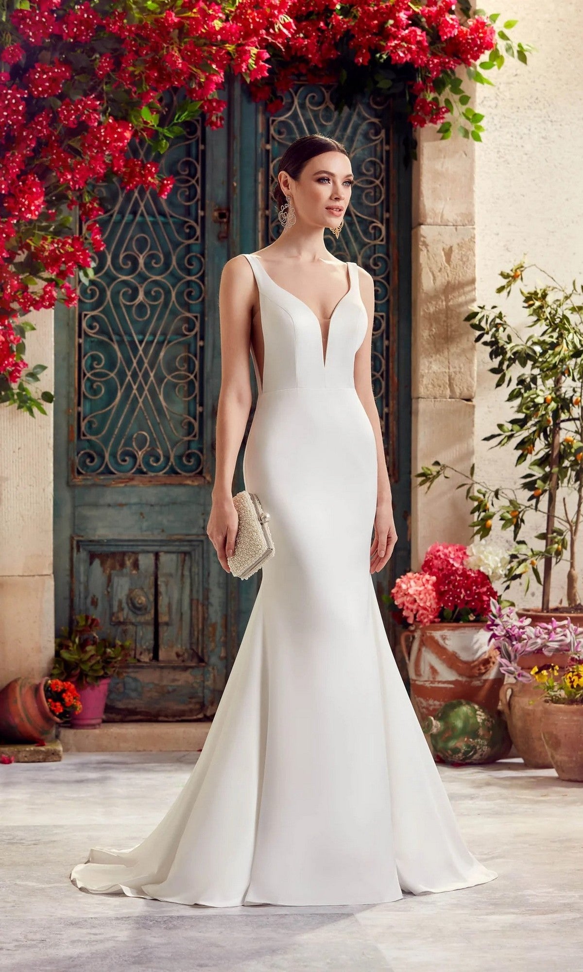 Wearing the Alyce 7052 long dress in diamond white, a woman stands outside by a green ornate door, surrounded by potted flowers and blooming bougainvillea, capturing modern bridal elegance.